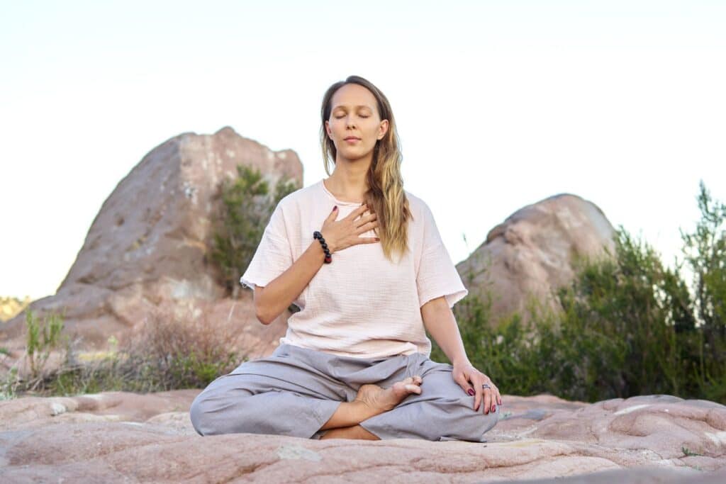 Blonde Frau in leichter Baumwollkleidung meditiert mit Hand auf dem Herzen auf einem Felsen